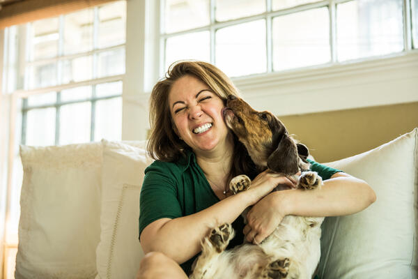 Woman sitting on her couch laughing while being licked by her dog