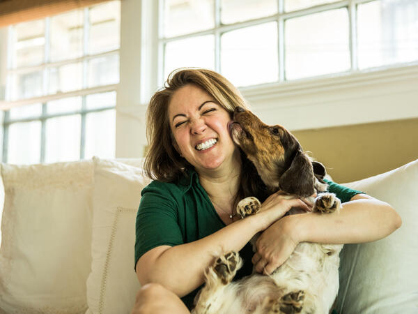 Woman sitting on her couch laughing while being licked by her dog