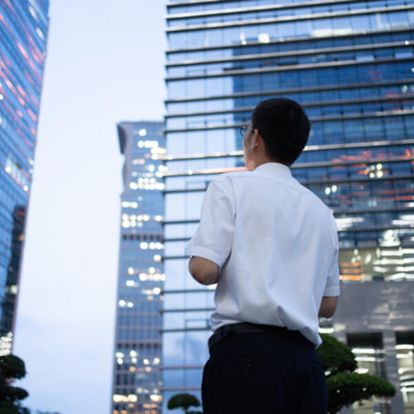 A young Asian business man in a white shirt looks up at tall buildings